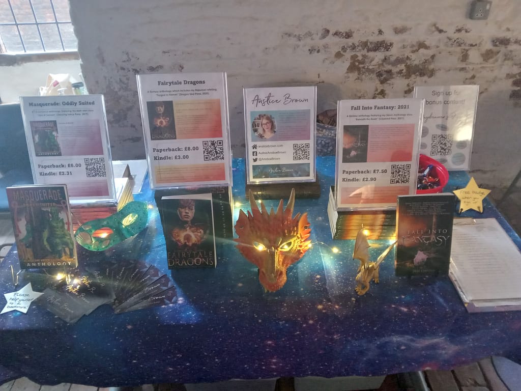 Anstice's anthologies on display at Boston Book Fest 2022 with an orange dragon mask and fairy lights on a galaxy print table cloth .