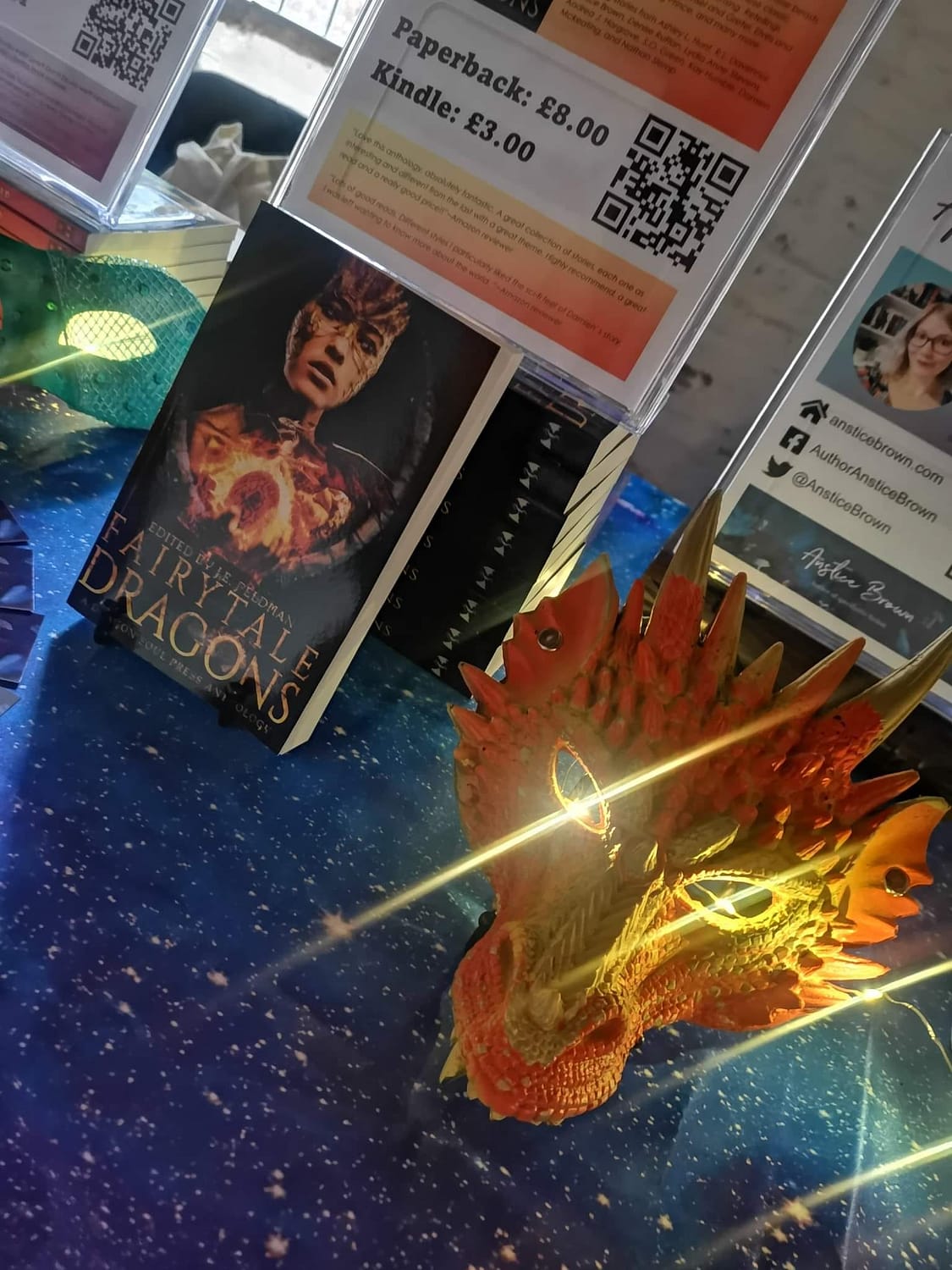 Fairytale Dragons anthology on display at Boston Book Fest 2022 with an orange dragon mask and fairy lights on a galaxy print table cloth 