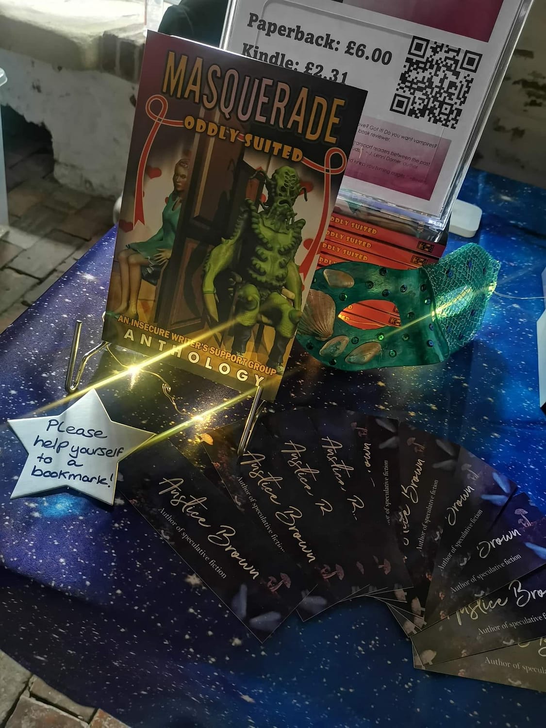 Masquerade: Oddly Suitedanthology on display at Boston Book Fest 2022 with an orangemermaid mask and fairy lights on a galaxy print table cloth