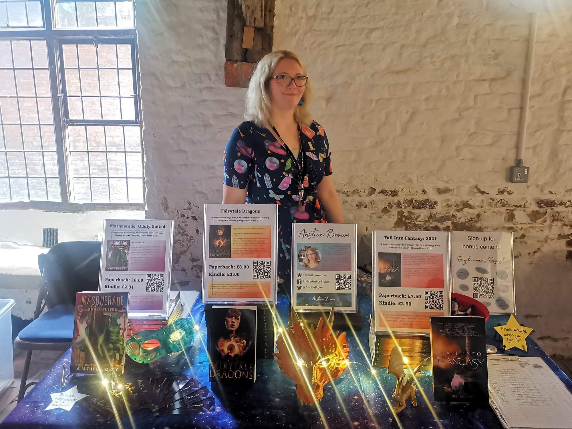 Author Anstice Brown wearing a potions and spell book print dress. She is smiling and standing behind her book stall displaying her anthologies. The stall is covered with a galaxy print tablecloth and decorated with fairy lights and props such as masquerade masks and dragon figurines.
