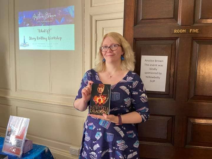 Author Anstice Brown smiles while holding her Fairytale Dragons anthollogy. She is wearing a blue book print dress and standing in front of a sign which reads "This event is sponsored by YellowBelly SUP"