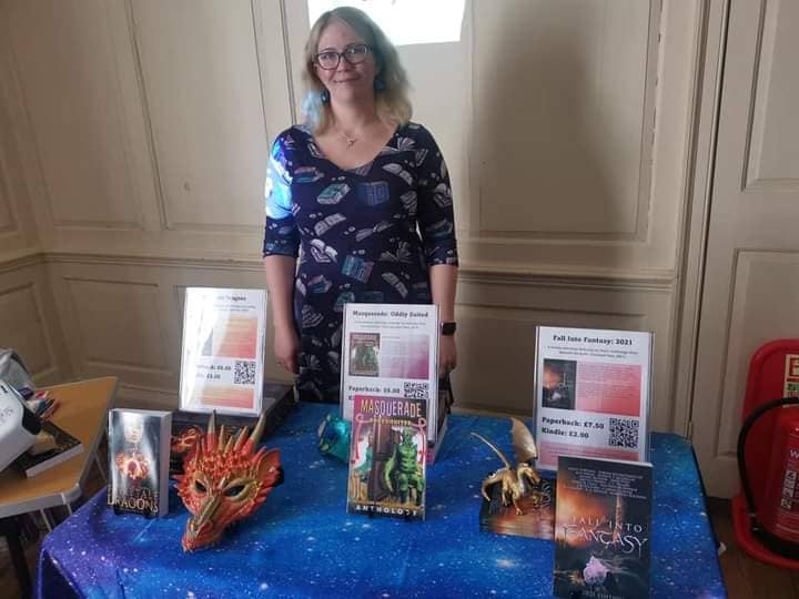 Author Anstice Brown standing behind her book stall. She is wearing a blue book print dress and smiling.