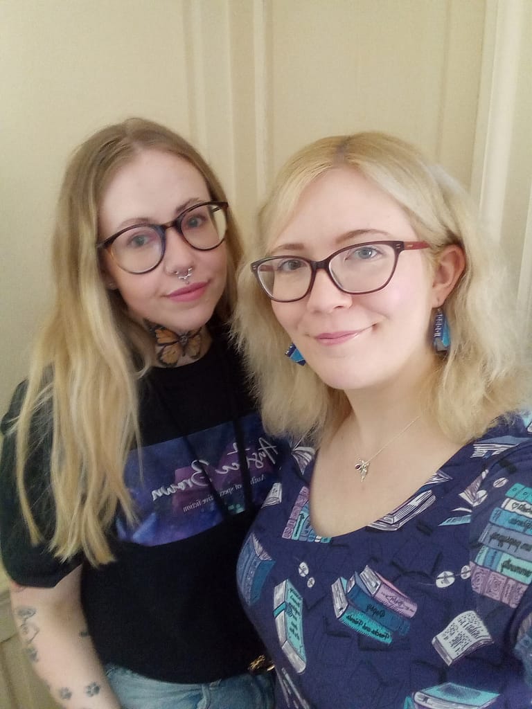 Author Anstice Brown and her sister Shana-Lee. Anstice is wearing a blue book print dress and Shana-Lee is wearing a T-shirt with Anstice's logo printed on it. Both have blonde hair and glasses and similar smiles.